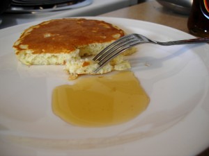 half a fluffy pancake and maple syrup on a plate, with a fork resting tines-down on a bite of pancake