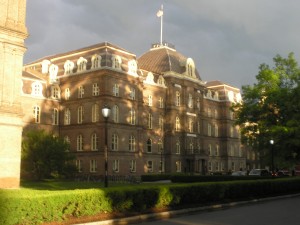 _big brick building in the sunlight, framed by dark thunderclouds and bright bits of green foliage_