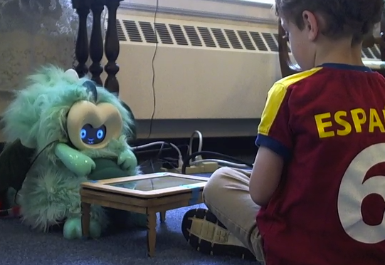 Boy sitting on the floor across a mini table from a dragon robot, looking at the robot intently